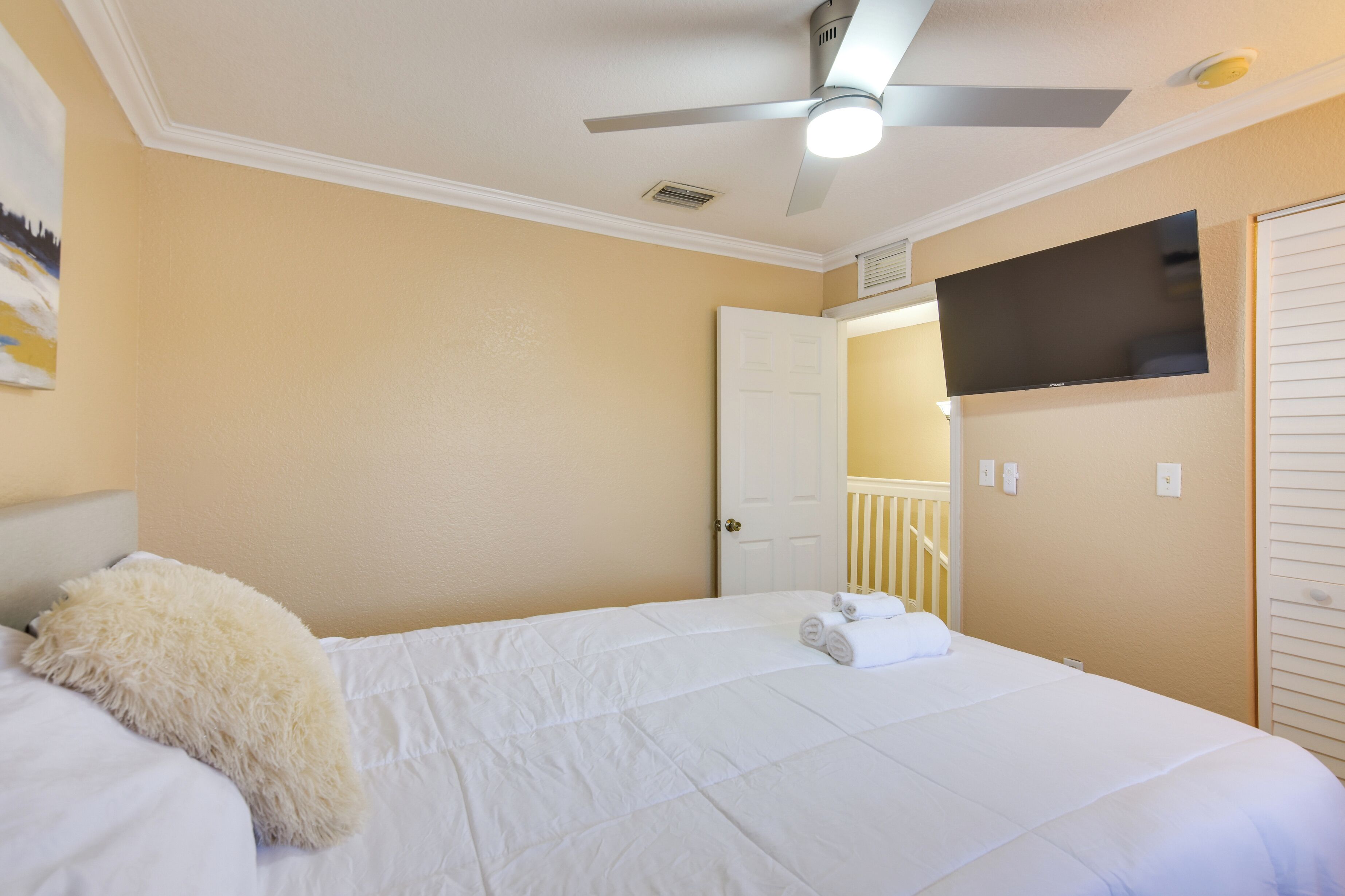 Une chambre à coucher comprenant un lit, un ventilateur de plafond, un téléviseur fixé au mur et une porte donnant sur une autre pièce.