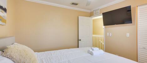 Une chambre à coucher comprenant un lit, un ventilateur de plafond, un téléviseur fixé au mur et une porte donnant sur une autre pièce.