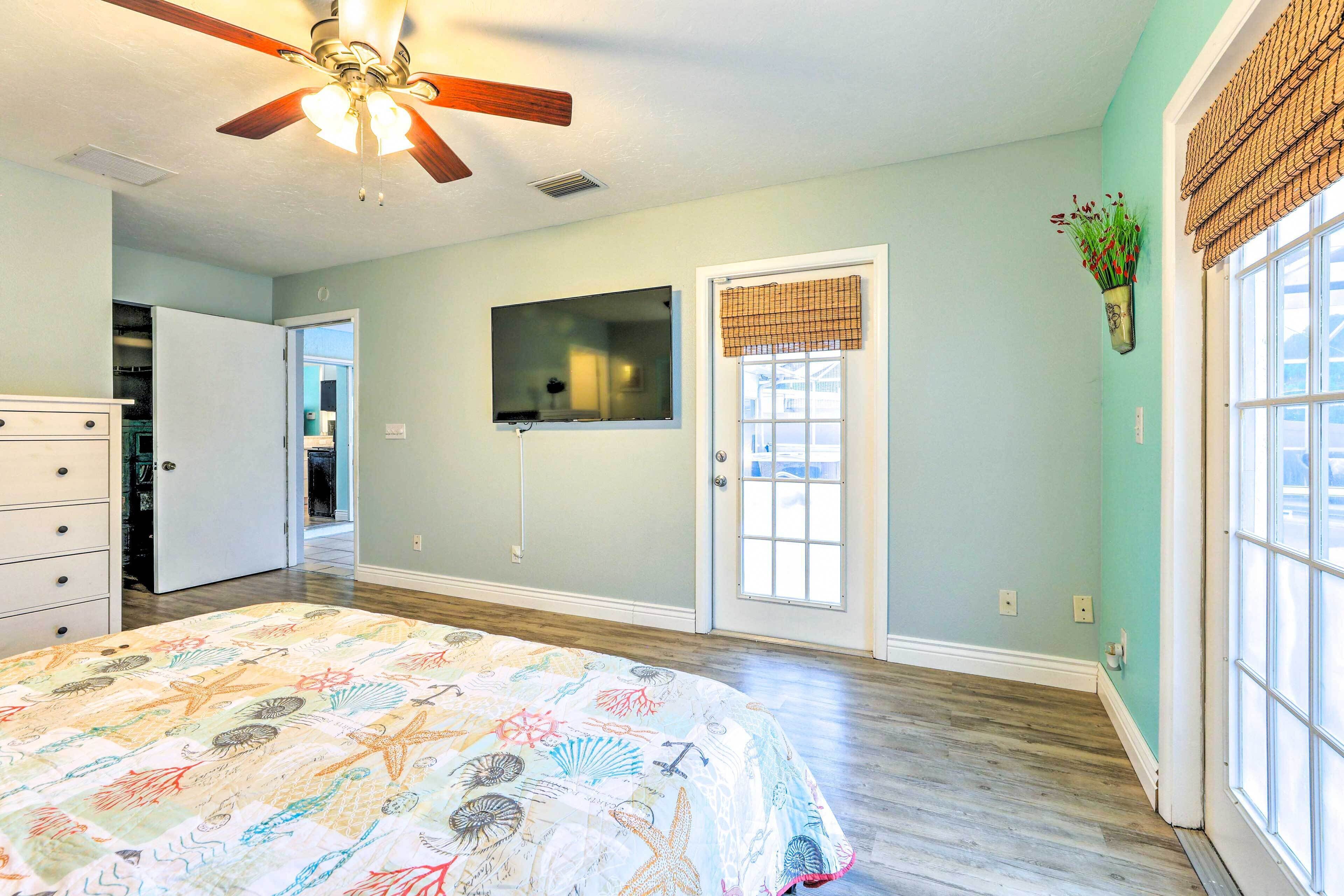 A bedroom with a bed, a ceiling fan, a door with a window, and a wall-mounted TV.