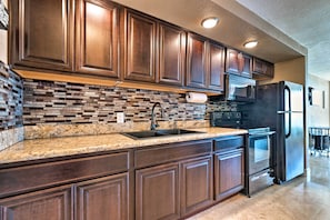 A kitchen with dark wooden cabinets, a granite countertop, a built-in brick backsplash, a stainless steel refrigerator, and a microwave. - Oceanfront Daytona Beach Studio w/ Balcony! (Daytona Beach Shores)