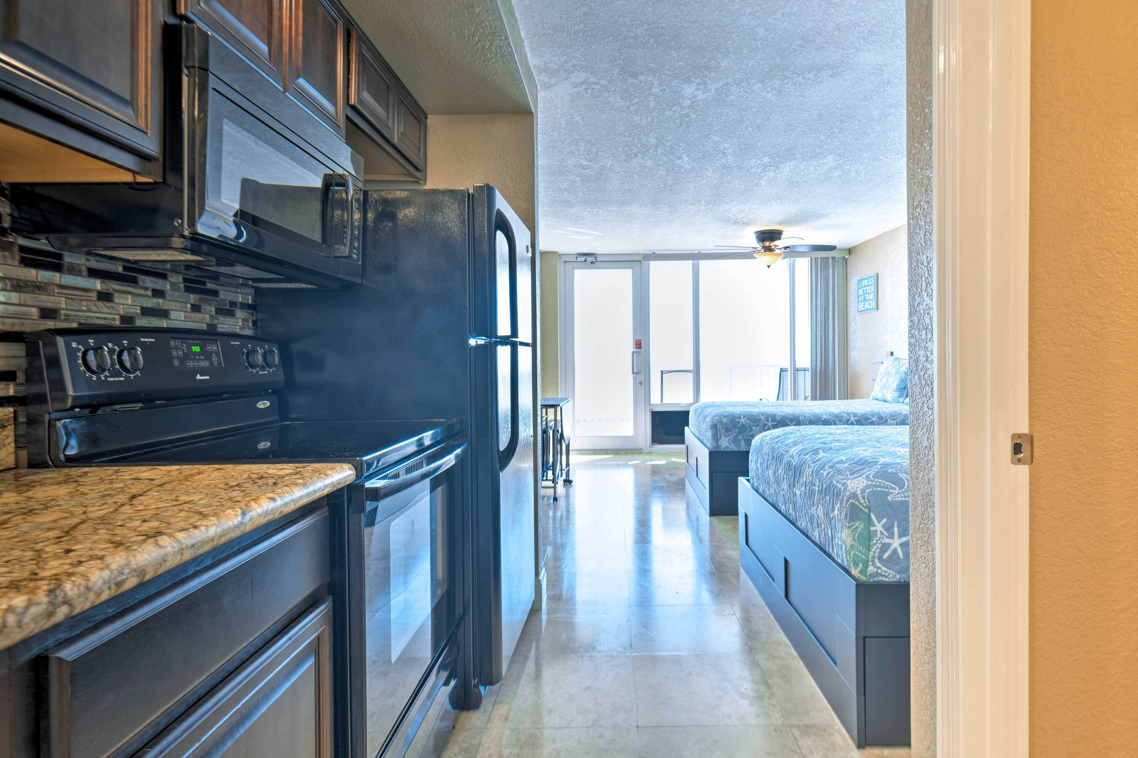 A compact kitchen with a stove, microwave, and refrigerator. A bed with a patterned bedspread is visible in the adjacent room.
