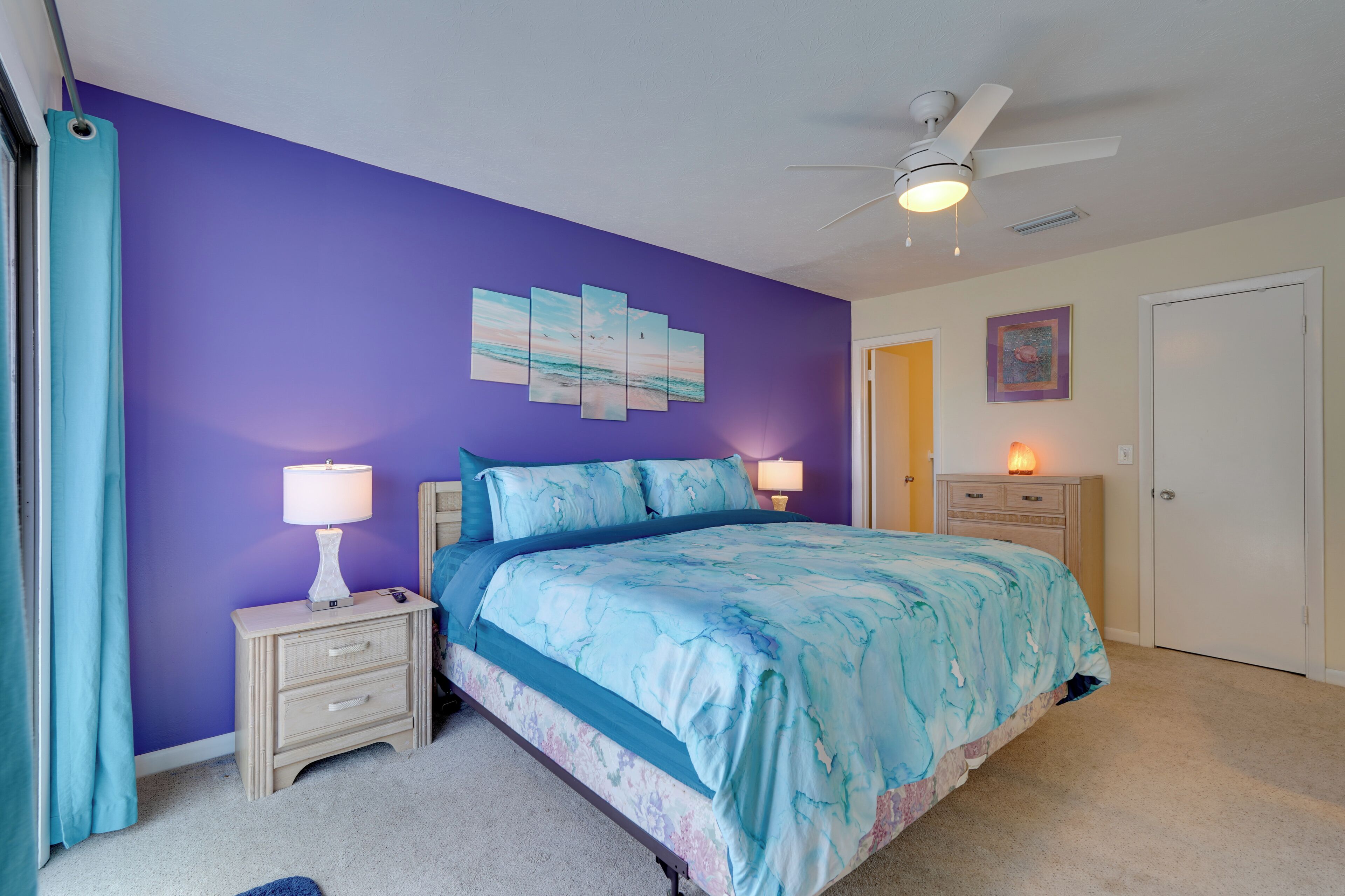 A bedroom with a purple wall, a bed with a blue and white bedspread, a nightstand with a lamp, and a ceiling fan.
