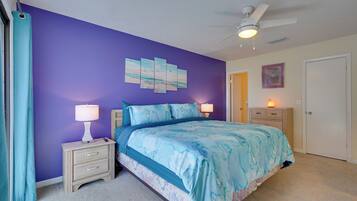 A bedroom with a purple wall, a bed with a blue and white bedspread, a nightstand with a lamp, and a ceiling fan.
