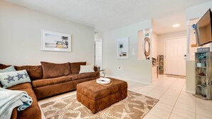 A living room with a brown sofa, a brown ottoman, and a patterned rug. There are framed pictures on the wall and a door labeled "BEACH HOUSE" to the right. - Pet-friendly Home w/ Pool in Palm Harbor! (Palm Harbor)