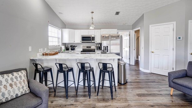 A modern kitchen with white cabinets, a central island with bar stools, and a stainless steel trash can. - Private Pool & Hot Tub at Panama City Home! (Panama City Beach)