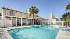 A swimming pool with a white fence, palm trees, and a building with a balcony in the background. - Private Pool & Hot Tub at Panama City Home! (Panama City Beach)