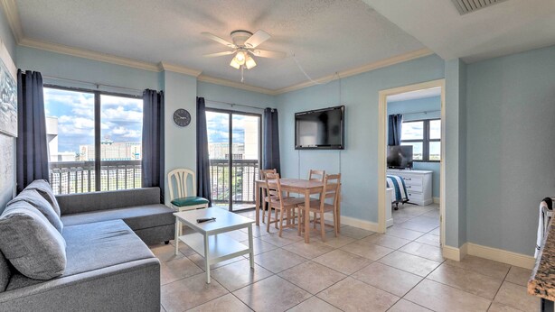 A living room with a sofa, a dining table with chairs, a ceiling fan, and a TV mounted on the wall.