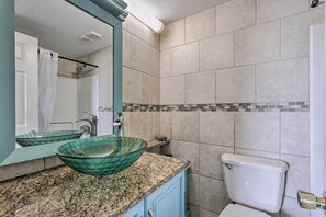 Une salle de bain dotée d’un comptoir en granit, d’un lavabo en verre, de toilettes blanches et d’un miroir avec un cadre de couleur turquoise.