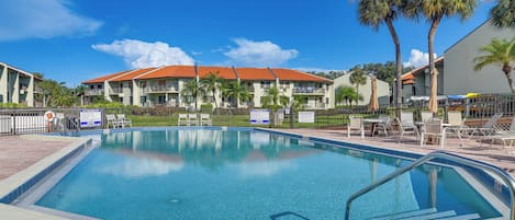 A swimming pool with a clear blue water surface, surrounded by lounge chairs and a building with a red-tiled roof.