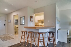 A modern kitchen with a wooden bar counter and stools, a refrigerator, and a dining area with a rug and chairs. - Ocean Views + Pool Access: Tierra Verde Condo! (Tierra Verde)
