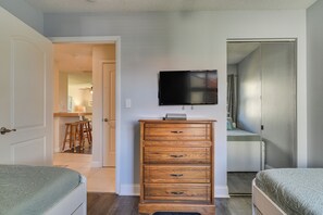 A bedroom with two beds, a wooden dresser, a flat-screen TV, and a door leading to a bathroom. - Ocean Views + Pool Access: Tierra Verde Condo! (Tierra Verde)