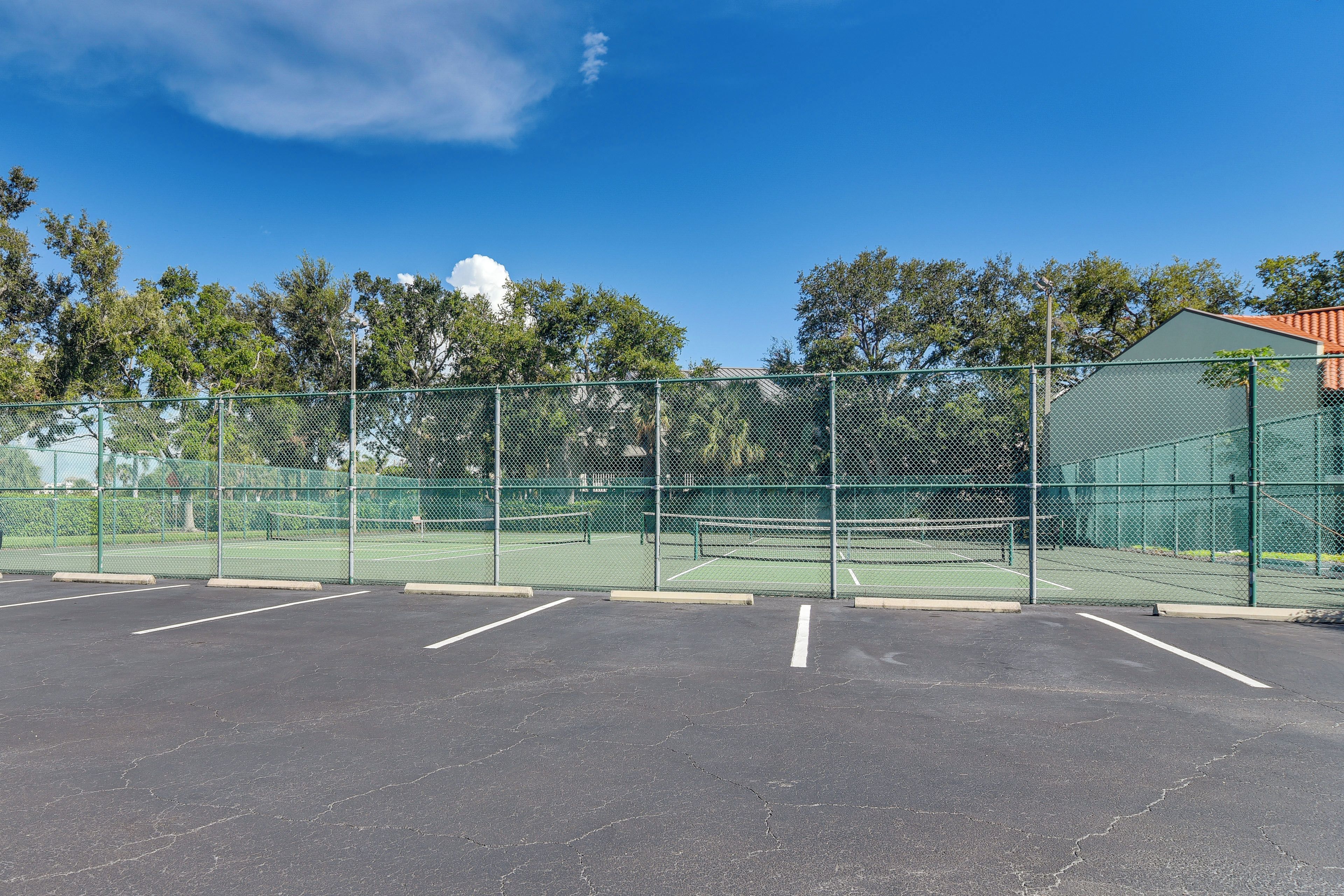Una cancha de tenis cercada con varios espacios de estacionamiento y un edificio verde al fondo.