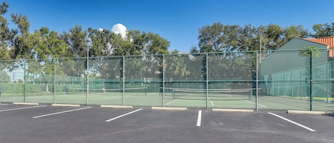 Una cancha de tenis cercada con varios espacios de estacionamiento y un edificio verde al fondo.