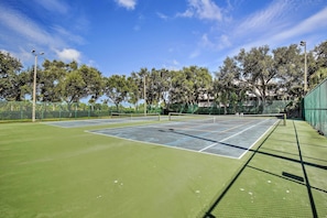Cancha de tenis al aire libre con red, rodeada de árboles y una valla.