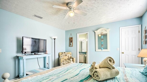 A bedroom with a bed, a television on a stand, a chair, a ceiling fan, and a door.