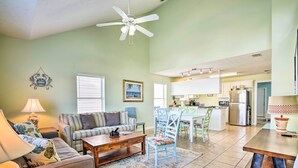 A spacious living room with a sofa, coffee table, and a dining area with chairs and a table. A ceiling fan is visible, and the kitchen area is in the background.