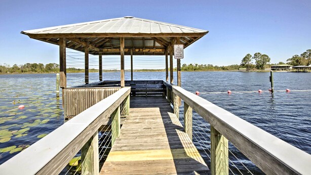 Un muelle de madera que conduce a un pabellón cubierto junto a un lago tranquilo con plantas flotantes.