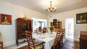 Un comedor con una mesa blanca dispuesta para seis personas, rodeada de sillas de madera con tapicería floral. Un candelabro cuelga del techo, y se aprecia un aparador de madera con cristalería en el fondo.