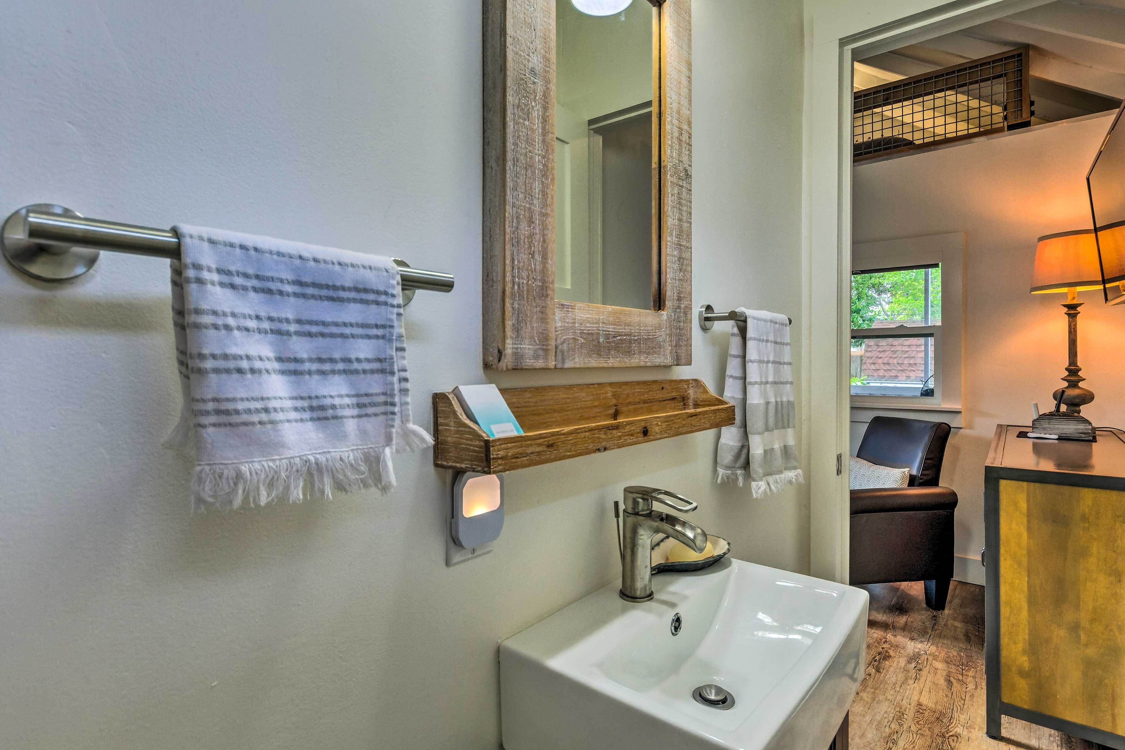 Une salle de bain avec un lavabo blanc, une étagère en bois sur laquelle est posée une serviette, un miroir et une chaise.