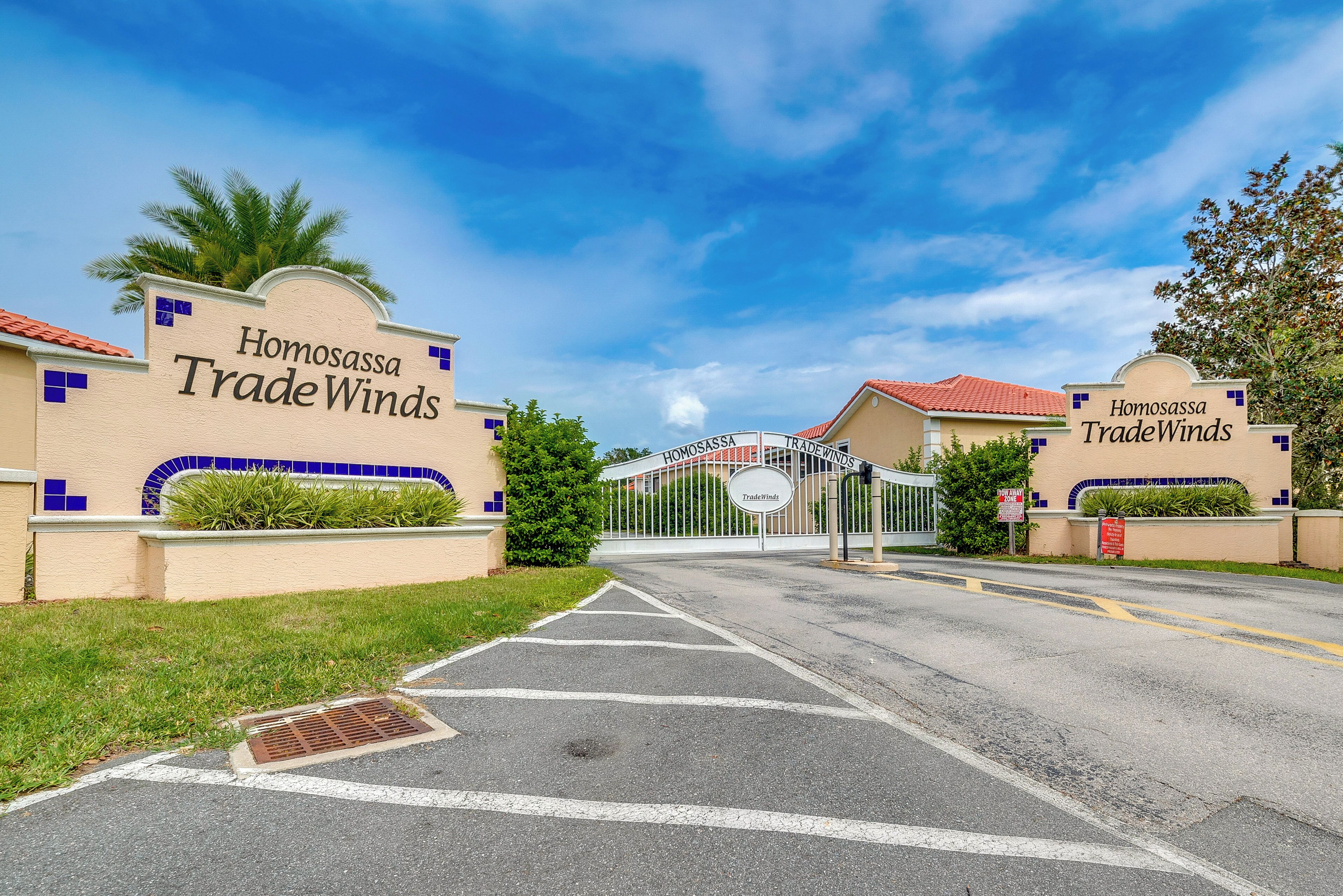 Una entrada con portón a Homosassa Trade Winds, con cielo despejado y palmeras.