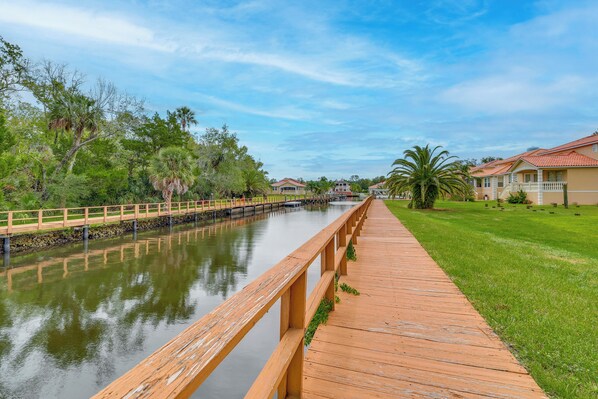 A wooden boardwalk along a calm water body with a bridge, surrounded by greenery and buildings with red roofs. - Resort Perks: Family Gem w/ Homosassa River Access (Homosassa)
