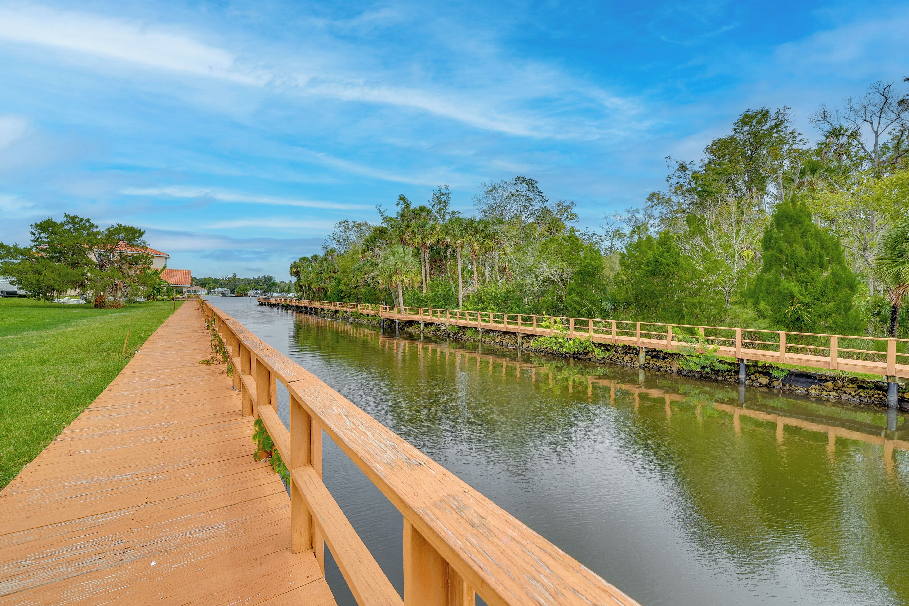 A wooden boardwalk along a calm river with a wooden bridge, surrounded by lush greenery and trees.