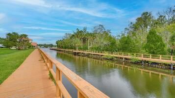 A wooden boardwalk along a calm river with a wooden bridge, surrounded by lush greenery and trees.
