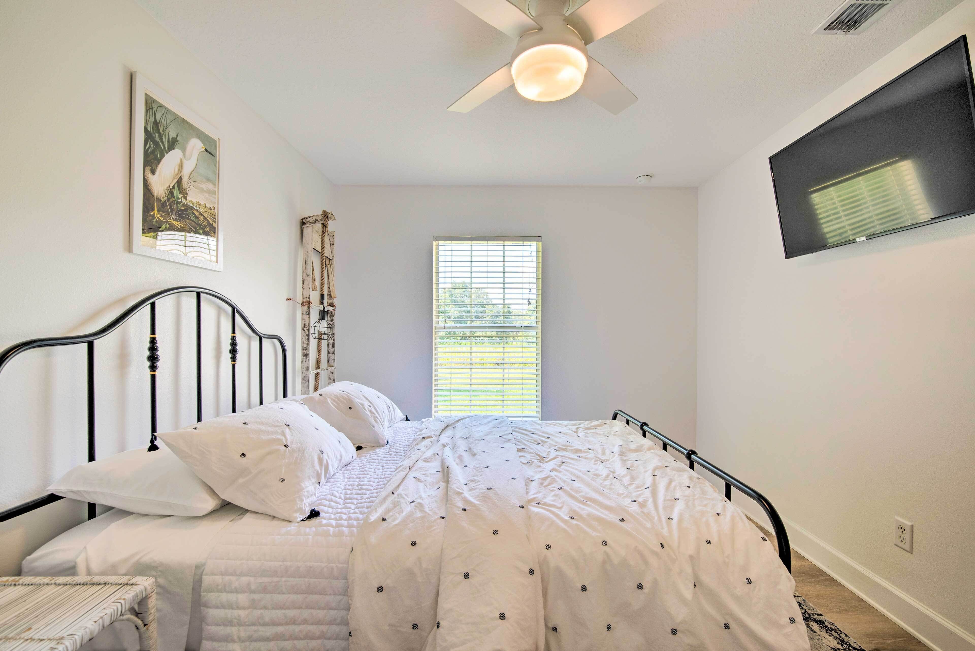 A bedroom with a bed, a ceiling fan, a window with blinds, and a wall-mounted TV.