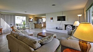 A living room with a sofa, a coffee table, and a lamp. There is a kitchen in the background with a TV and a fan. - Pet-friendly Daytona Beach Home, Walk to the Ocean (Port Orange)
