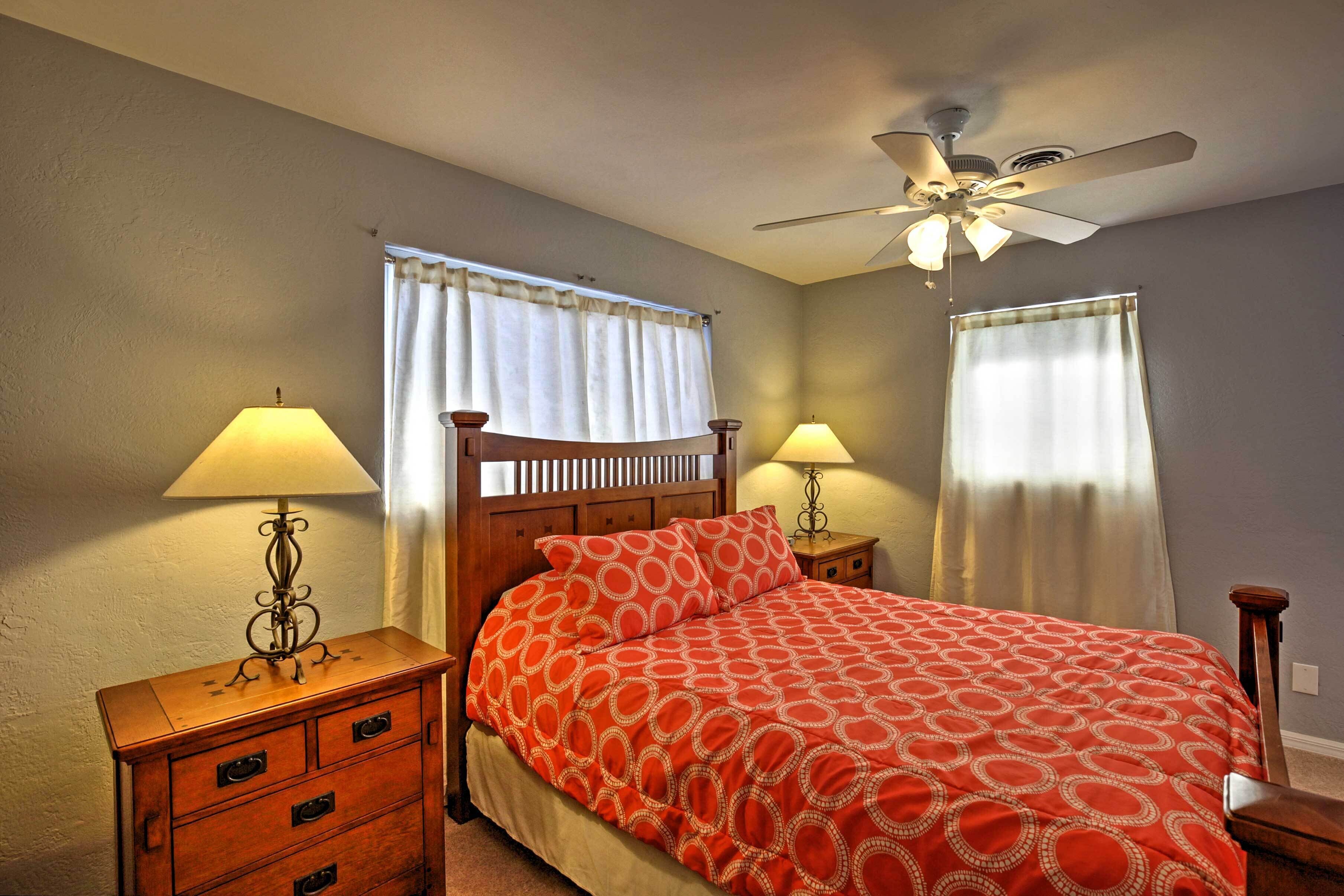 A bedroom with a bed, two bedside tables with lamps, a ceiling fan, and curtains on the windows.