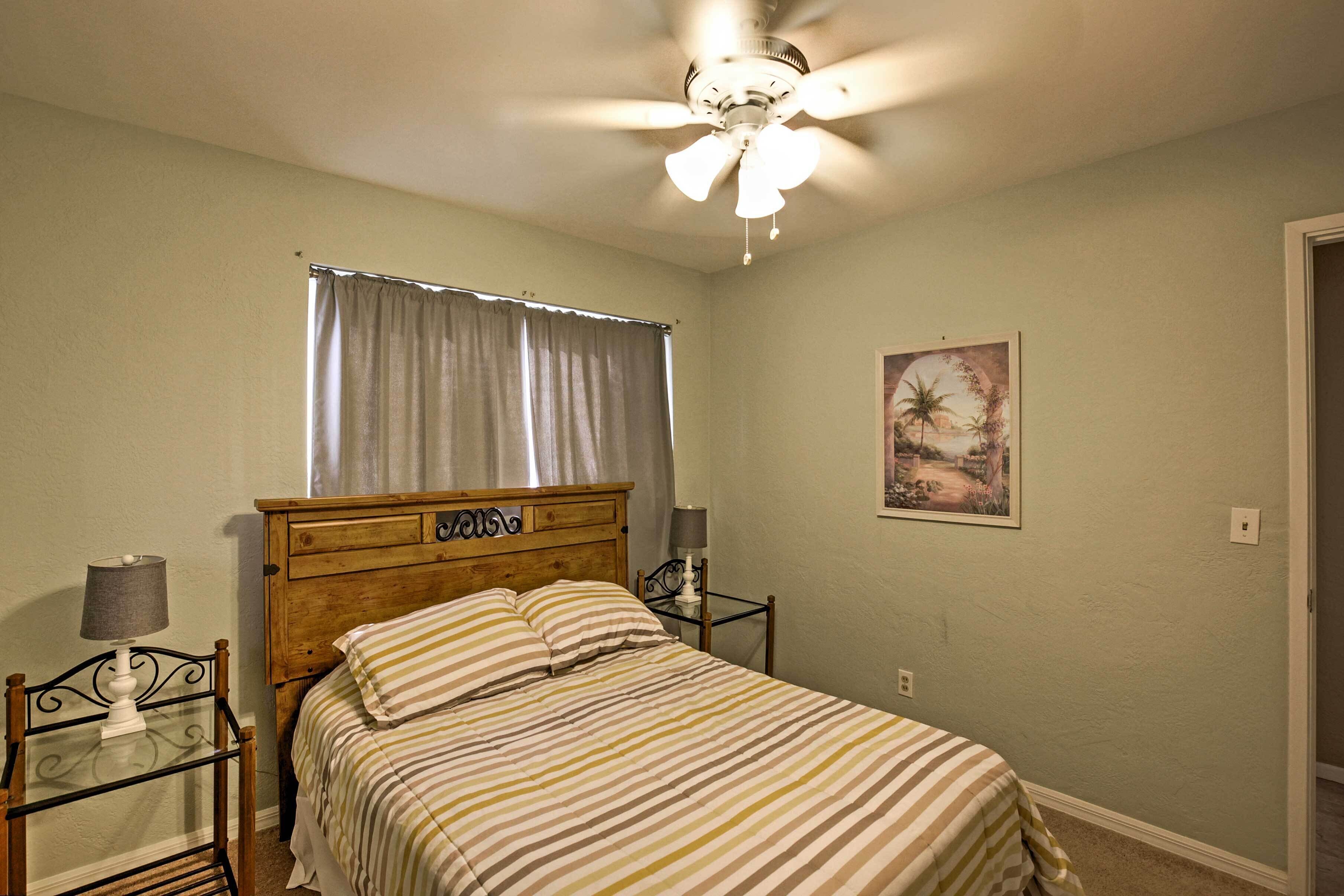 A bedroom with a bed, a ceiling fan, a nightstand, a lamp, a window with curtains, and a wall-mounted picture.