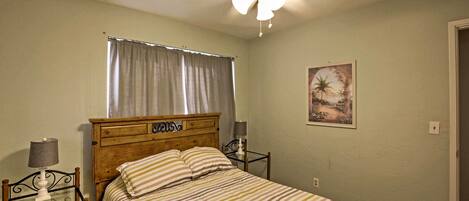 A bedroom with a bed, a ceiling fan, a nightstand, a lamp, a window with curtains, and a wall-mounted picture.