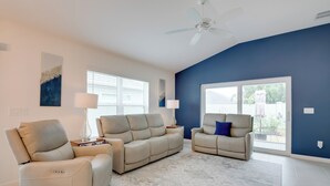 A spacious living room with a beige sectional sofa, a smaller armchair, and a coffee table. There are two floor lamps, a ceiling fan, and a large window providing natural light. - Lanai & Resort Amenities: The Villages Getaway (Wildwood)