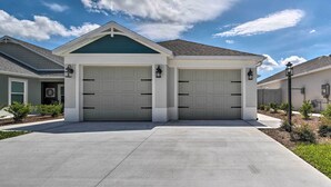A two-car garage with a green roof and white walls, a concrete driveway, and a clear blue sky with clouds. - Lanai & Resort Amenities: The Villages Getaway (Wildwood)