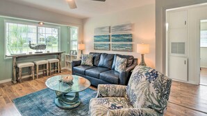 A living room with a black leather couch, a glass coffee table, and a floral armchair. There is a dining area with a bench and stools, and a ceiling fan above. - Punta Gorda Retreat Near Historic Downtown! (Punta Gorda)
