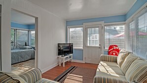 Una sala de estar con un sofá, un televisor sobre un mueble bajo, una puerta y una ventana con persianas.