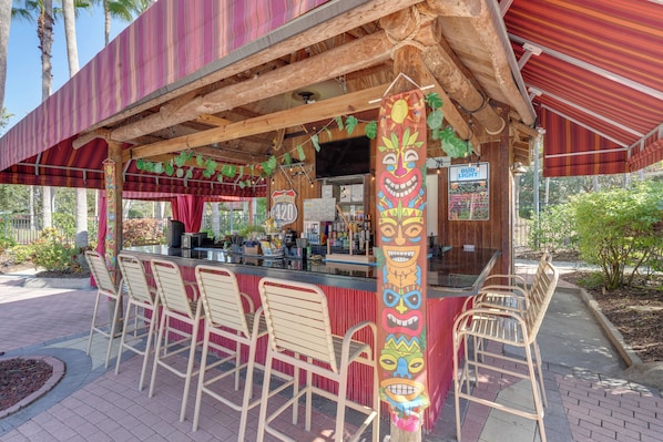 An outdoor bar with a tiki theme, featuring a counter, bar stools, and a wooden structure.