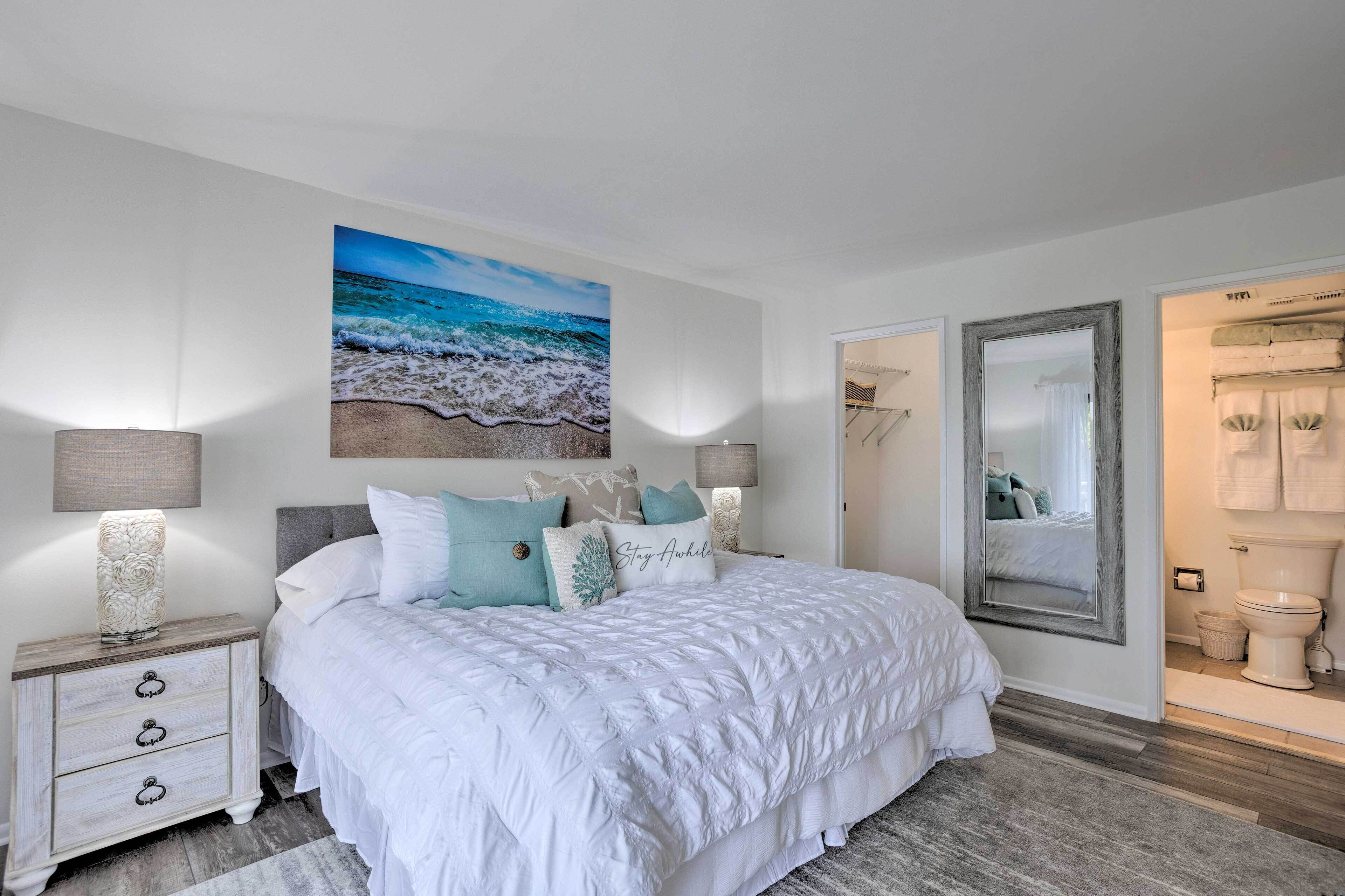 A bedroom with a large bed, white bedding, a beach-themed wall art, a nightstand with a lamp, and a mirror.