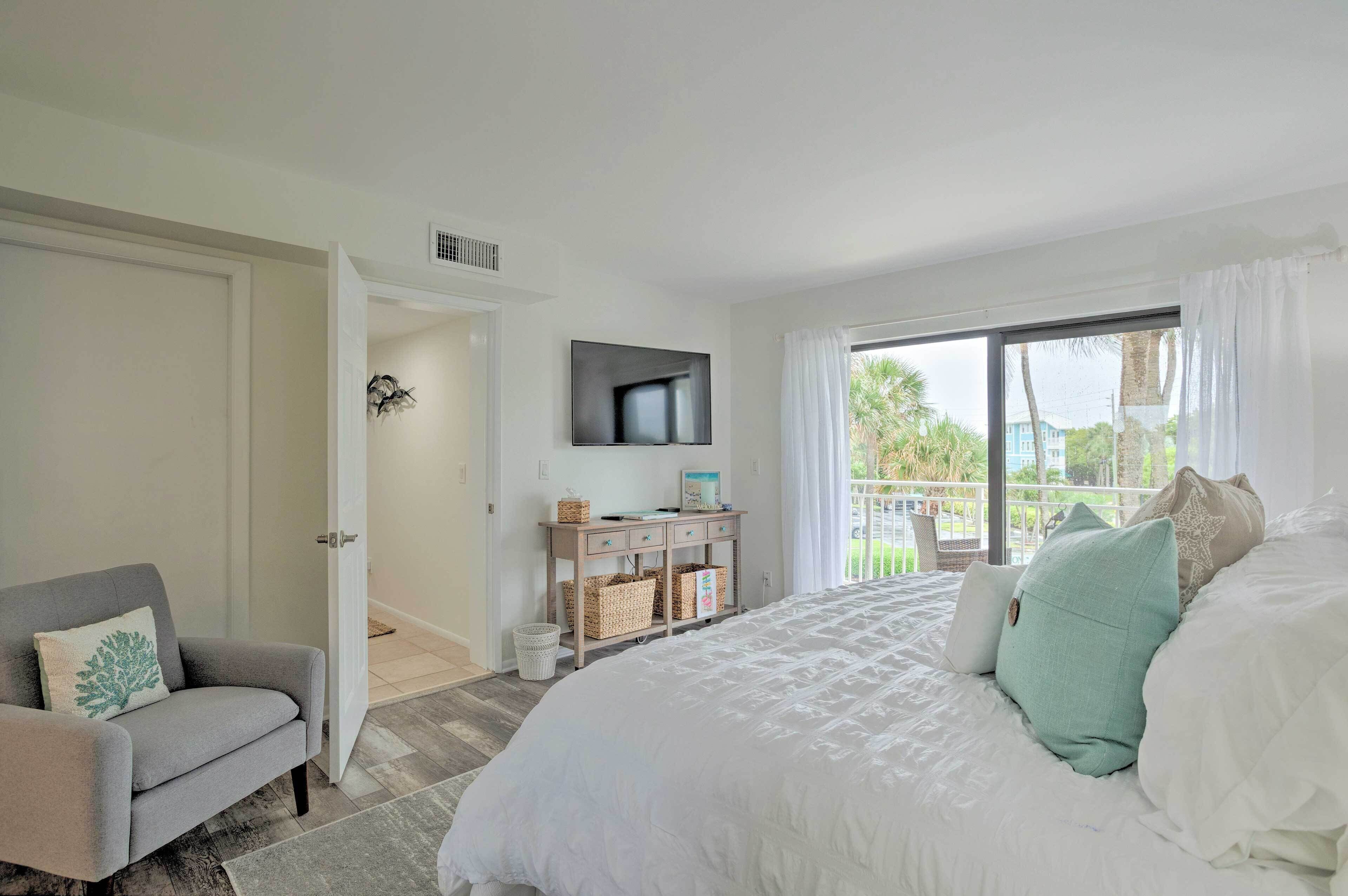 A bedroom with a large bed, a TV mounted on the wall, a desk with baskets, and a view of the outdoors through a window.