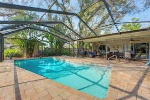 A covered swimming pool with a patio area, lounge chairs, and a view of trees.