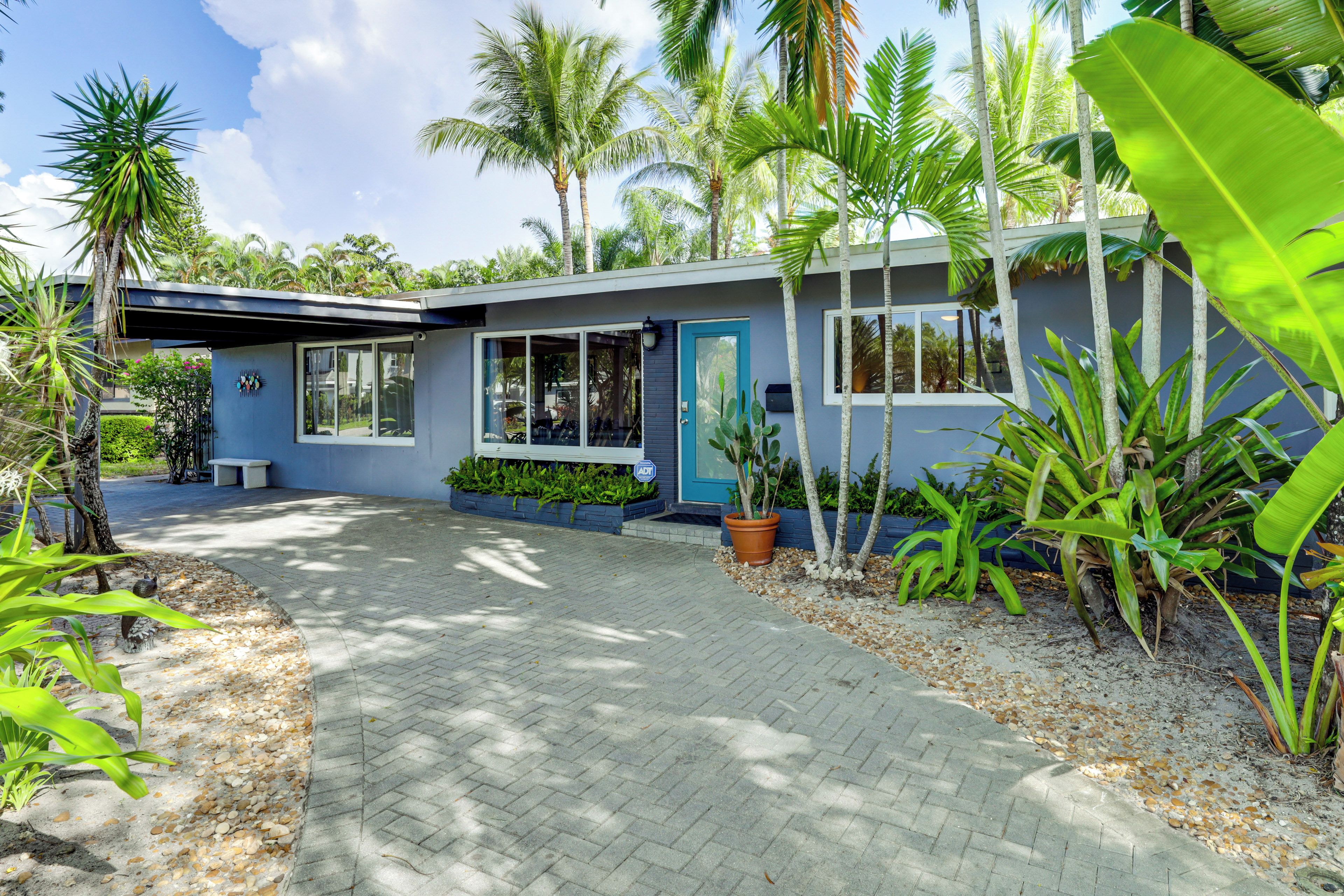 A modern house with a paved driveway, a blue door, and a garden with various plants.