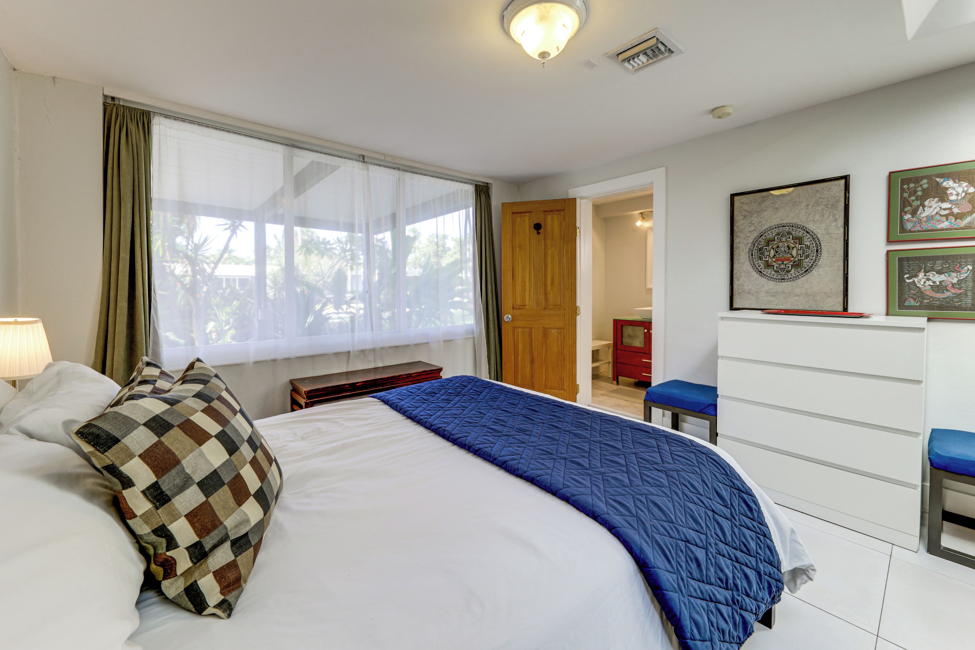 Une chambre à coucher comprenant un lit, une table de chevet, une fenêtre avec des rideaux, une porte, une commode et deux tableaux encadrés accrochés au mur.