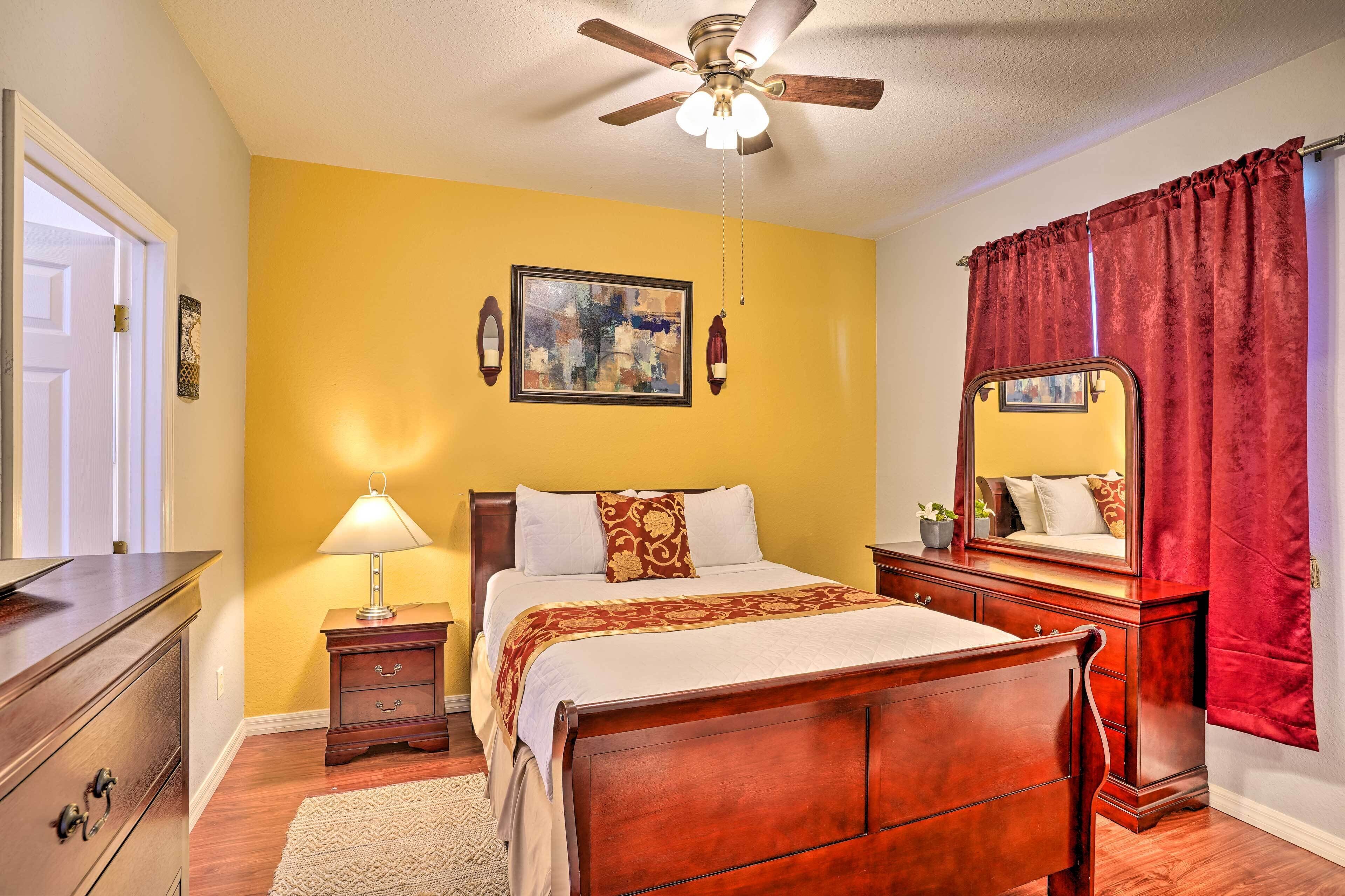 Une chambre à coucher comprenant un lit, une table de chevet, une lampe, une commode, un ventilateur de plafond et un grand miroir.