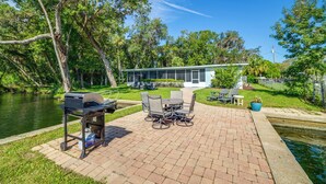 Eine Terrasse mit Grill, Esstisch und Stühlen, mit Blick auf ein Gewässer und ein Haus.