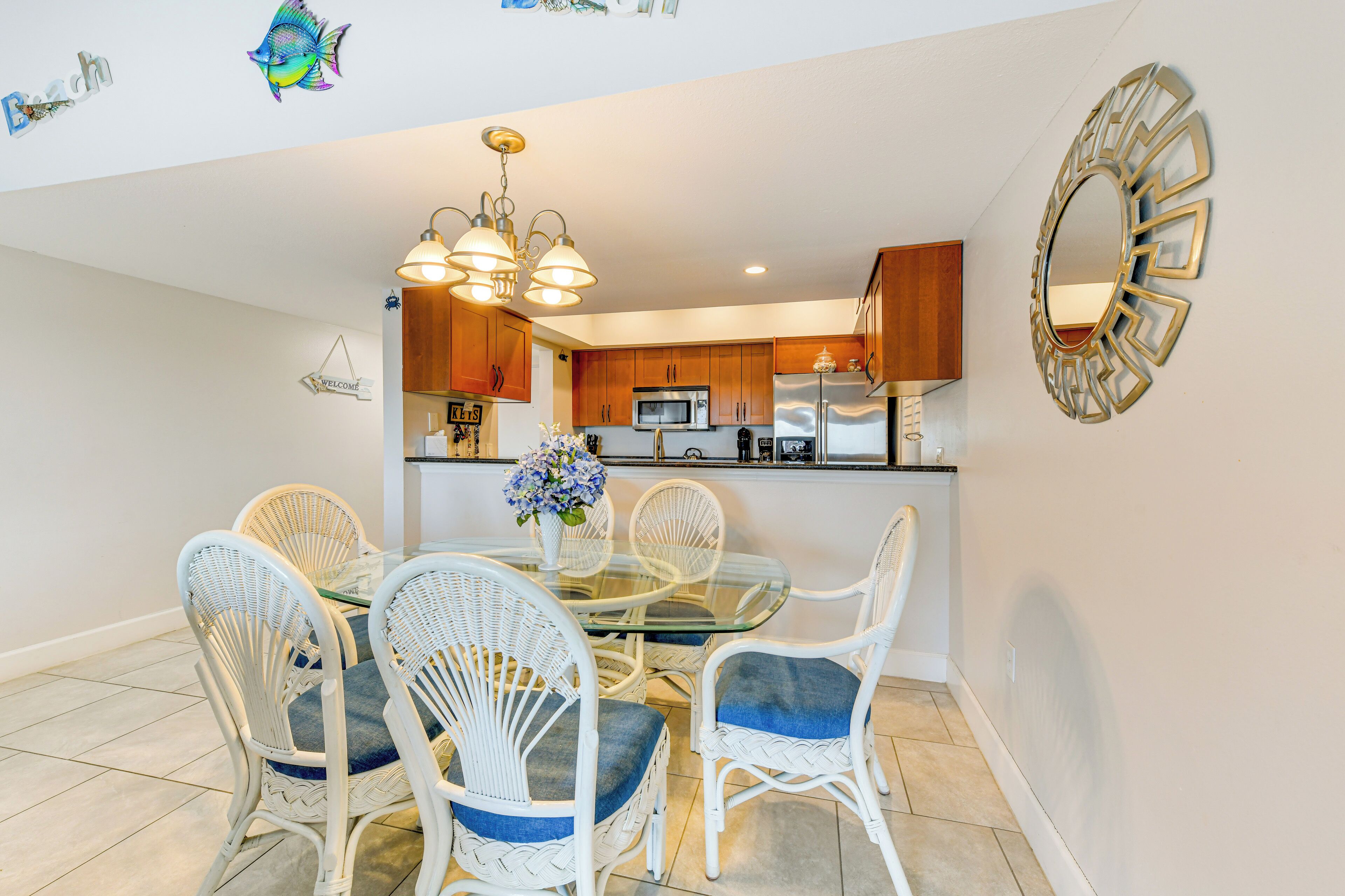 A dining area with a glass table and white wicker chairs, a kitchen in the background, and a decorative mirror on the wall.
