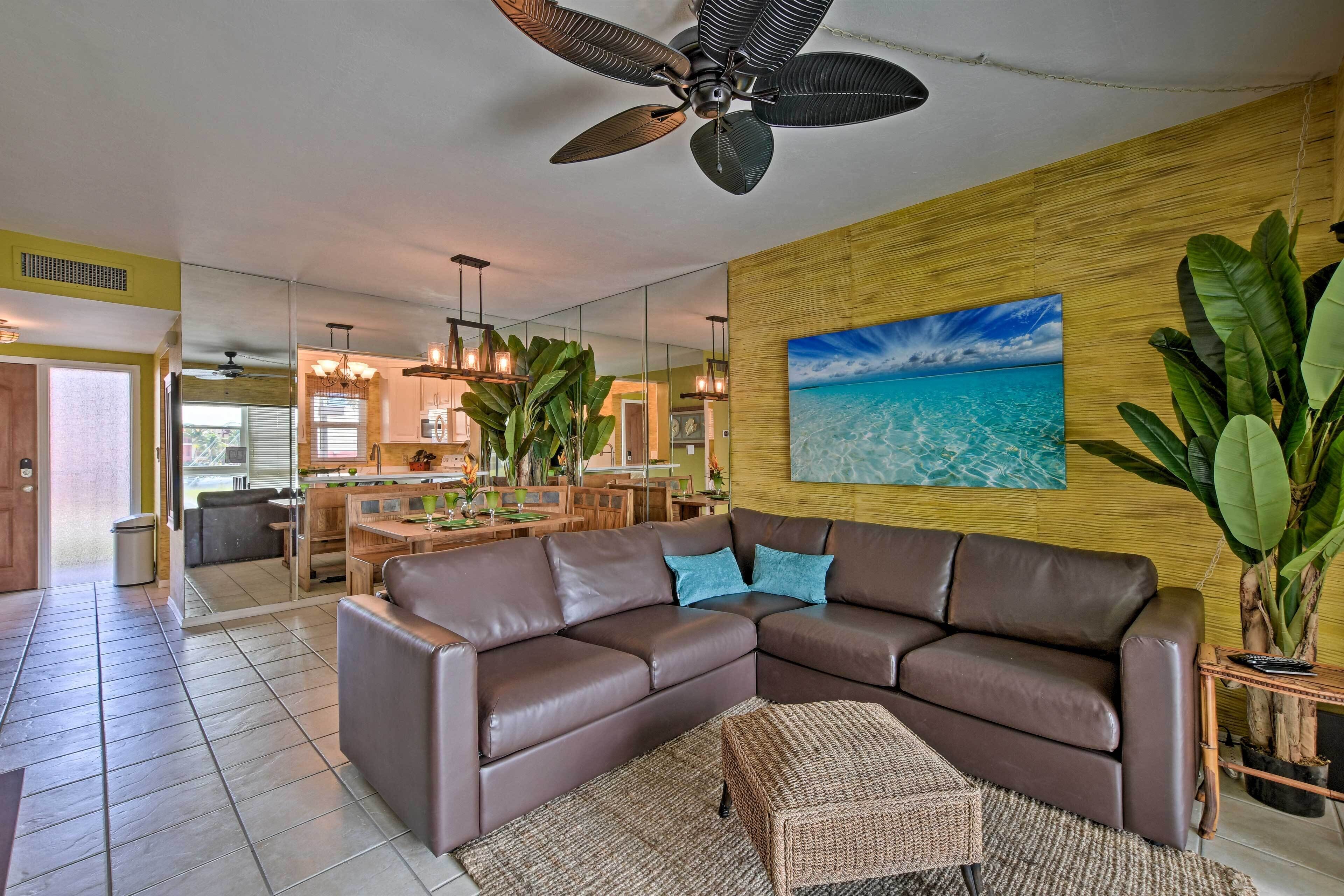Een moderne woonkamer met een hoekbank, een plafondventilator, een groot schilderij van een strand en een eethoek op de achtergrond.