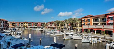 A marina with boats docked, surrounded by multi-story buildings with balconies and palm trees.