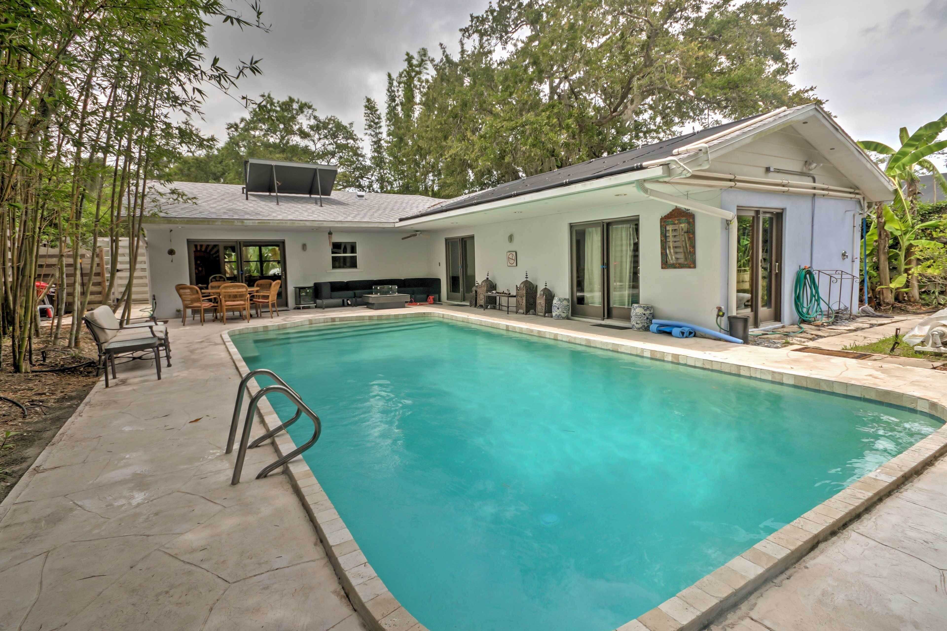 A rectangular swimming pool with clear blue water, surrounded by a concrete deck. There are wooden chairs and a table near the pool, and a covered patio area with a table and chairs.