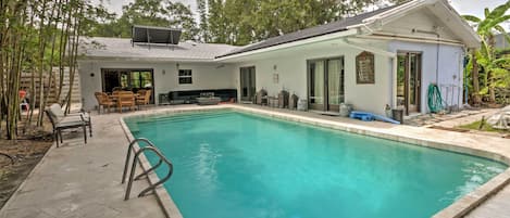 A rectangular swimming pool with clear blue water, surrounded by a concrete deck. There are wooden chairs and a table near the pool, and a covered patio area with a table and chairs.
