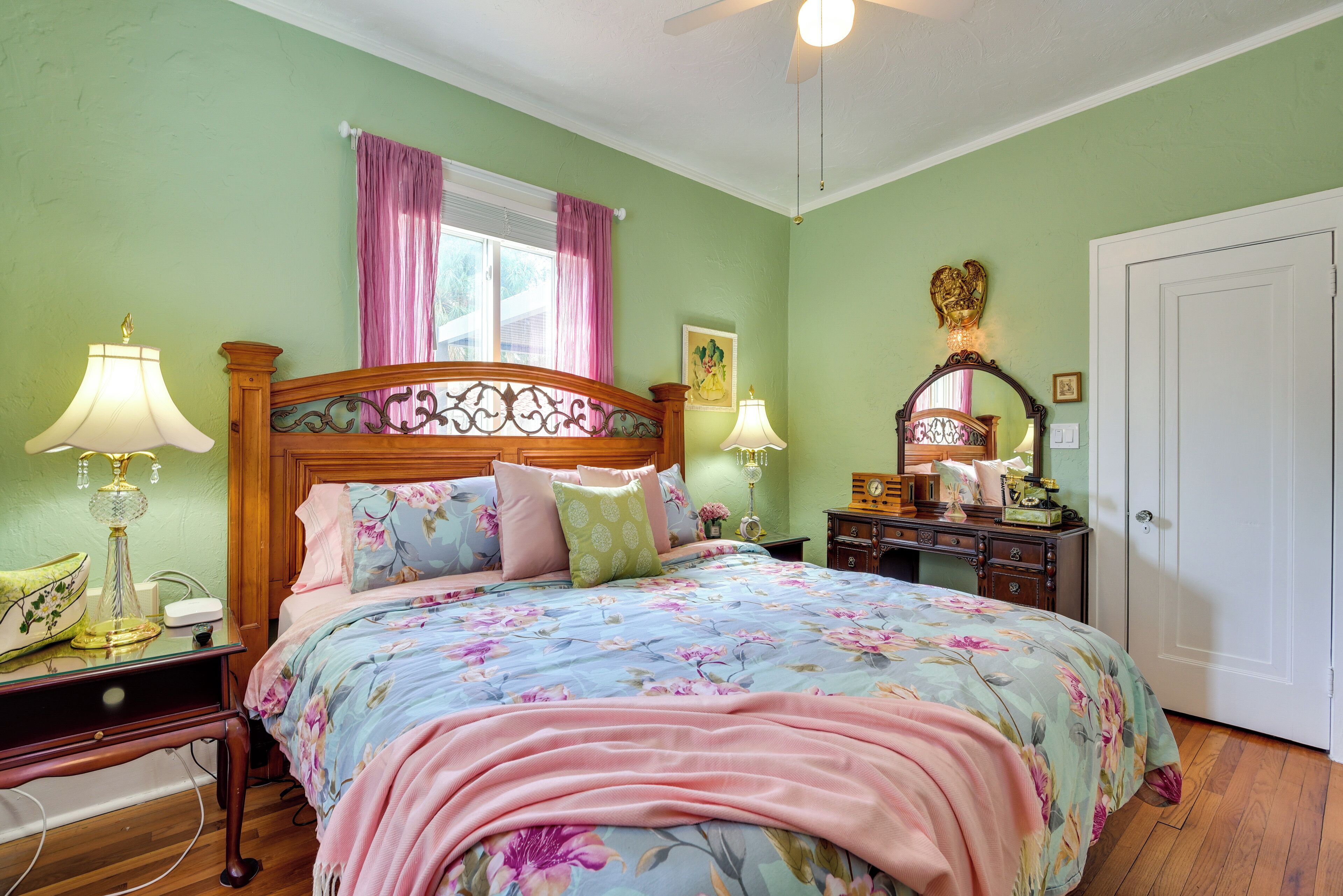 Une chambre à coucher avec une couvre-lit à motifs floraux, deux lampes de chevet, une tête de lit en bois, une fenêtre avec des rideaux roses et une porte blanche.